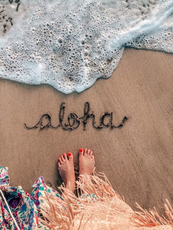 Shems feet with "aloha" written int he sand at the shoreline